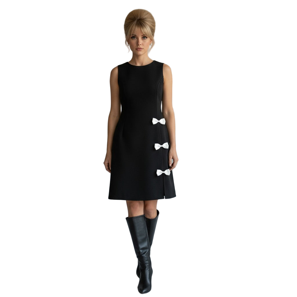 Woman wearing a black dress with white bow details and black knee-high boots on a plain background