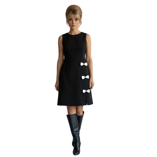 Woman wearing a black dress with white bow details and black knee-high boots on a plain background